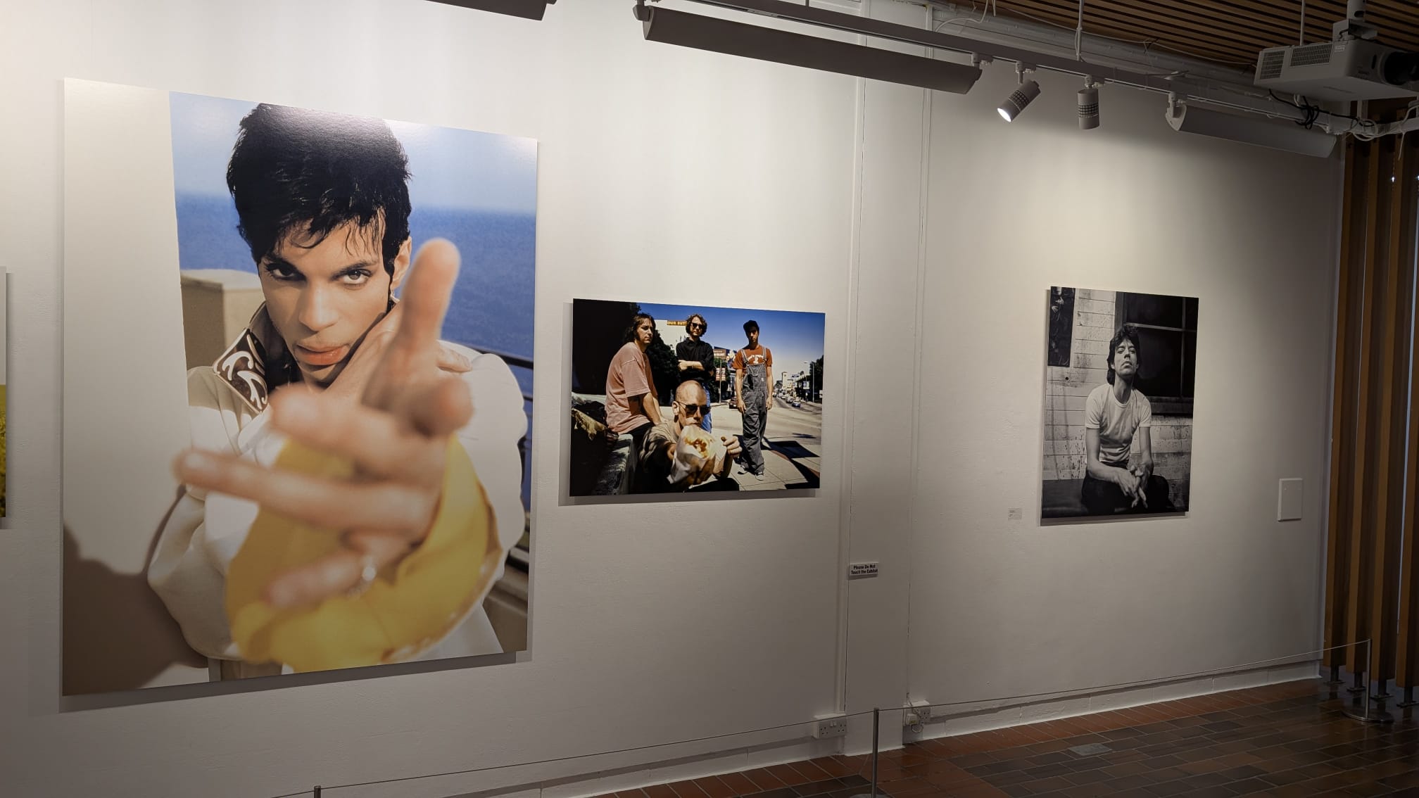 Three framed photographs hang on a gallery wall, including a large colour portrait on the left, a smaller group scene in the centre and a black-and-white portrait on the right.