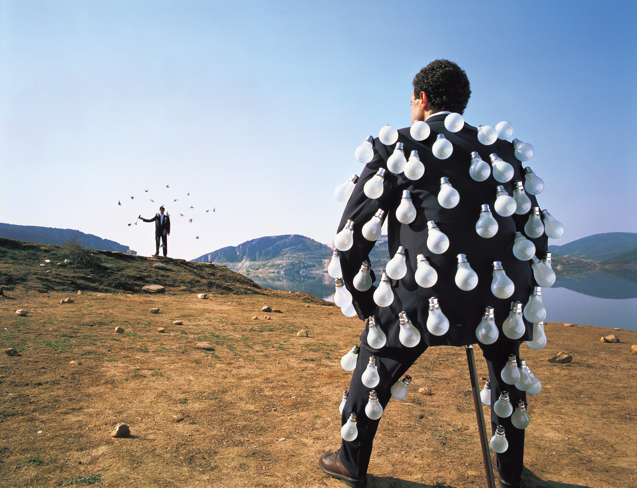 Person in black costume covered with white teardrop shapes stands on barren landscape, facing distant figure with arms raised near lake.