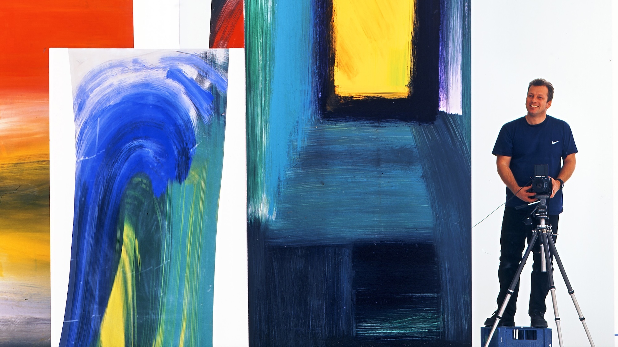 Man with tripod stands beside large abstract paintings featuring bold blue, yellow, orange and green brushstrokes and geometric shapes.