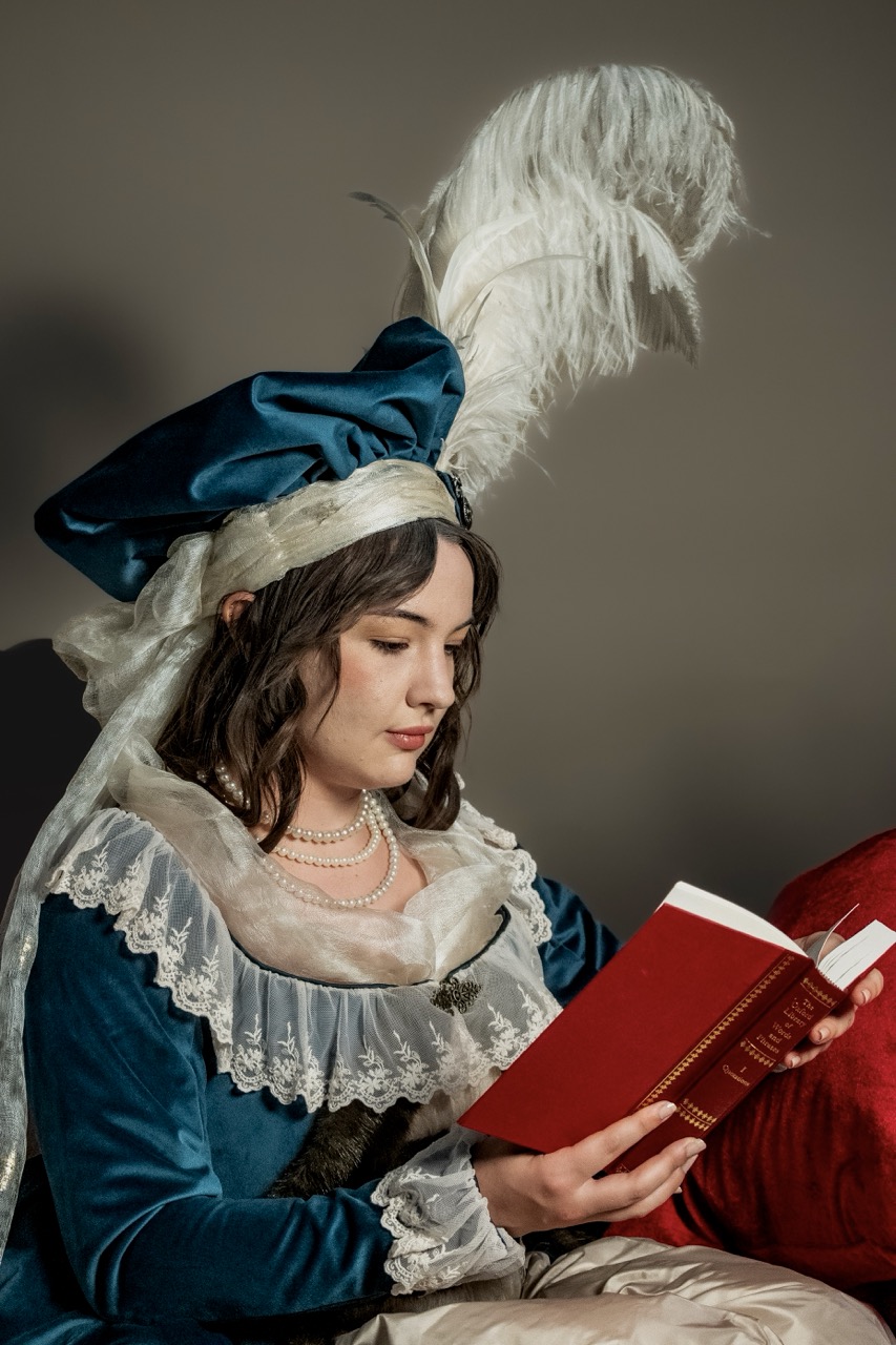 A woman in a blue velvet historical dress and feathered hat reads a red book, seated against a plain grey background.