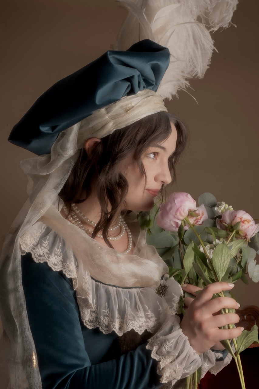 A woman in a teal historical dress and feathered bonnet holds a bouquet of pale pink flowers against a plain brown background.