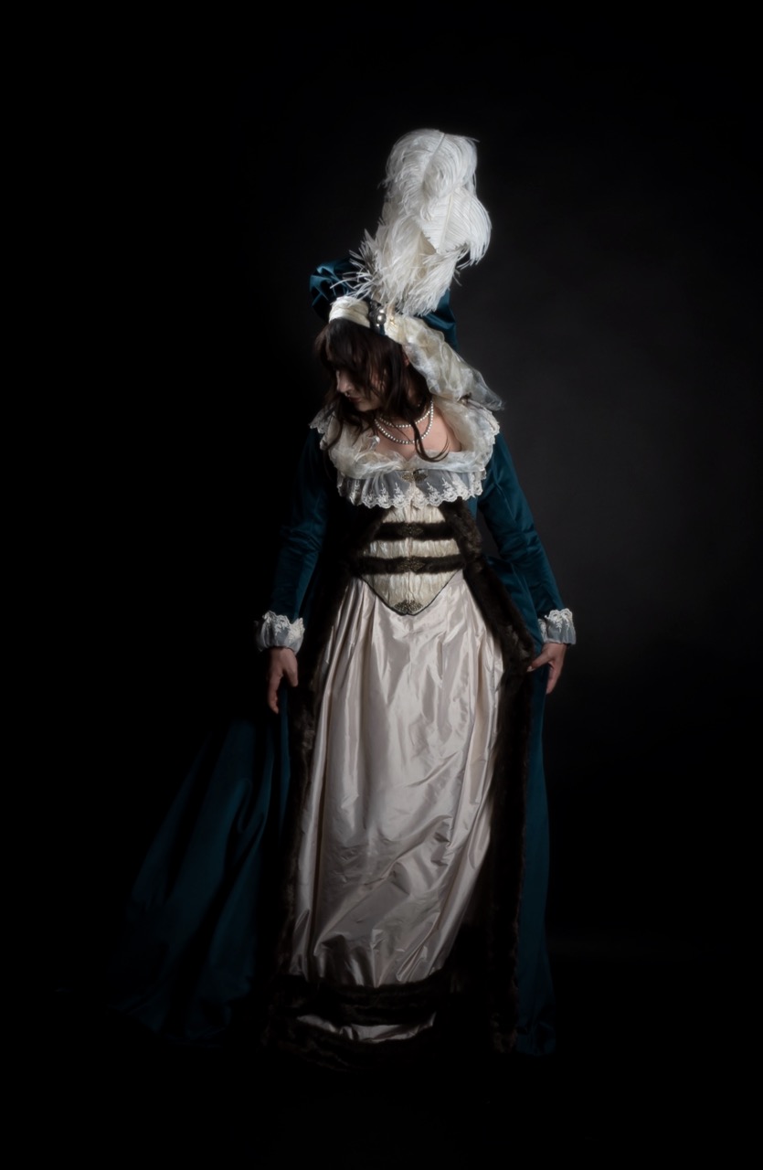 A woman in a blue and white historical costume with a feathered headdress stands against a black background.