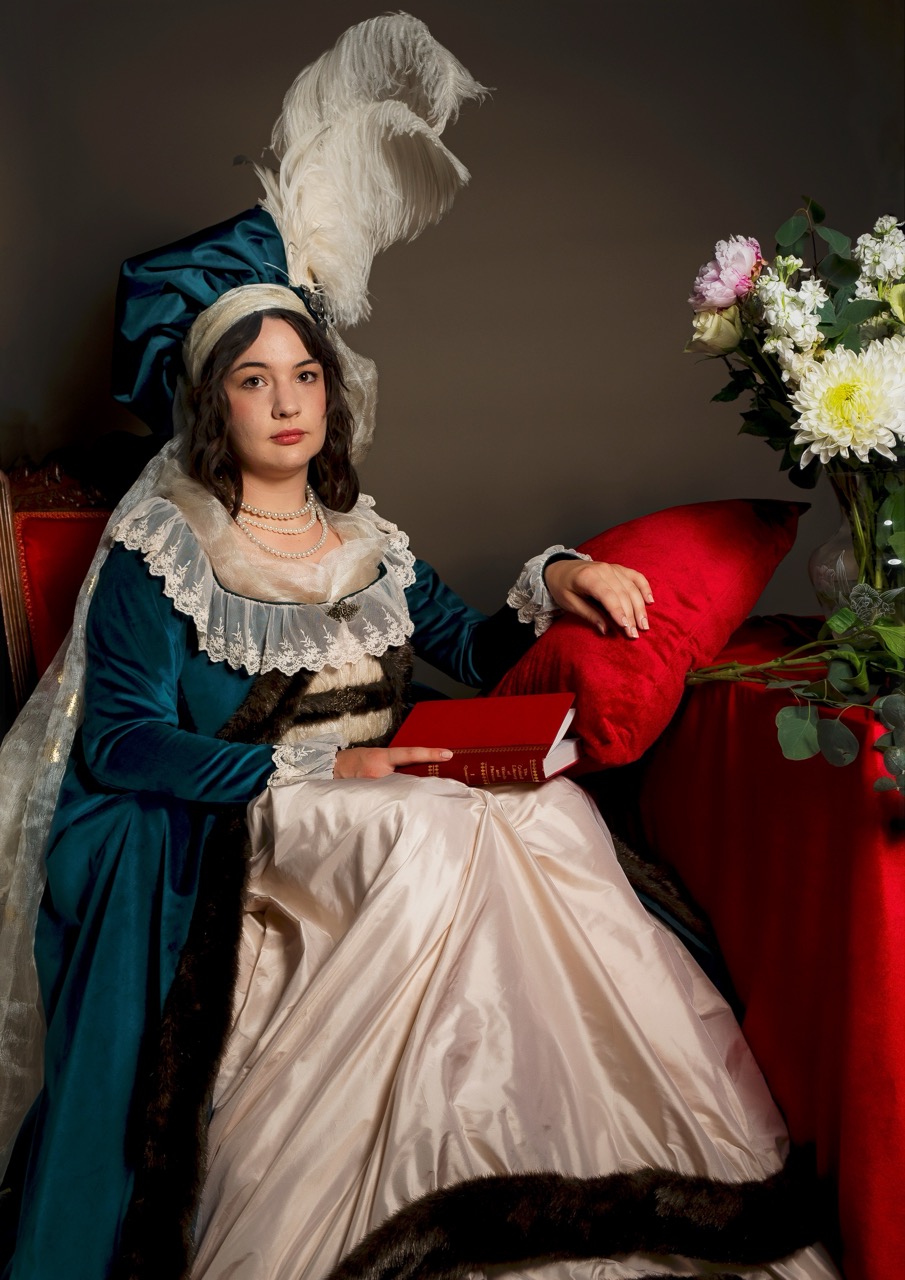 A woman in a blue historical dress and feathered headdress sits on a red chair holding a red book, with a vase of flowers beside her.