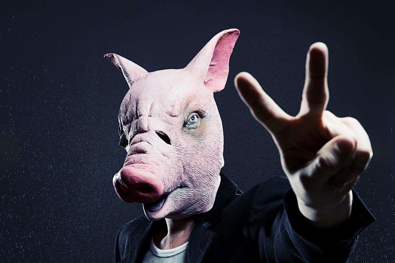 Person wearing pink pig mask with raised hand against dark background, dramatic lighting creating shadows.
