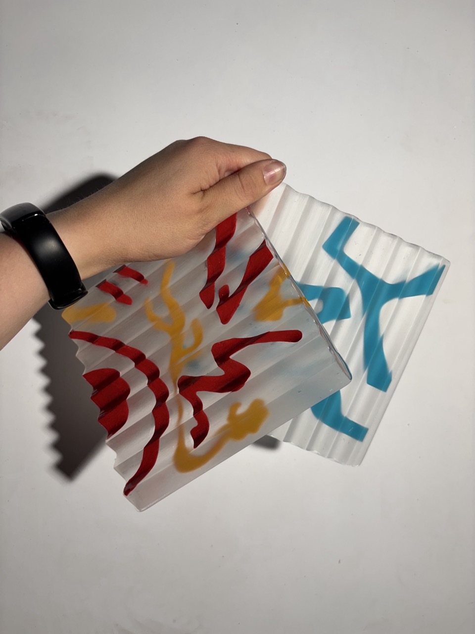 Hand holding white paper with red, yellow, and blue painted brushstrokes. Person wearing black fitness tracker on wrist.