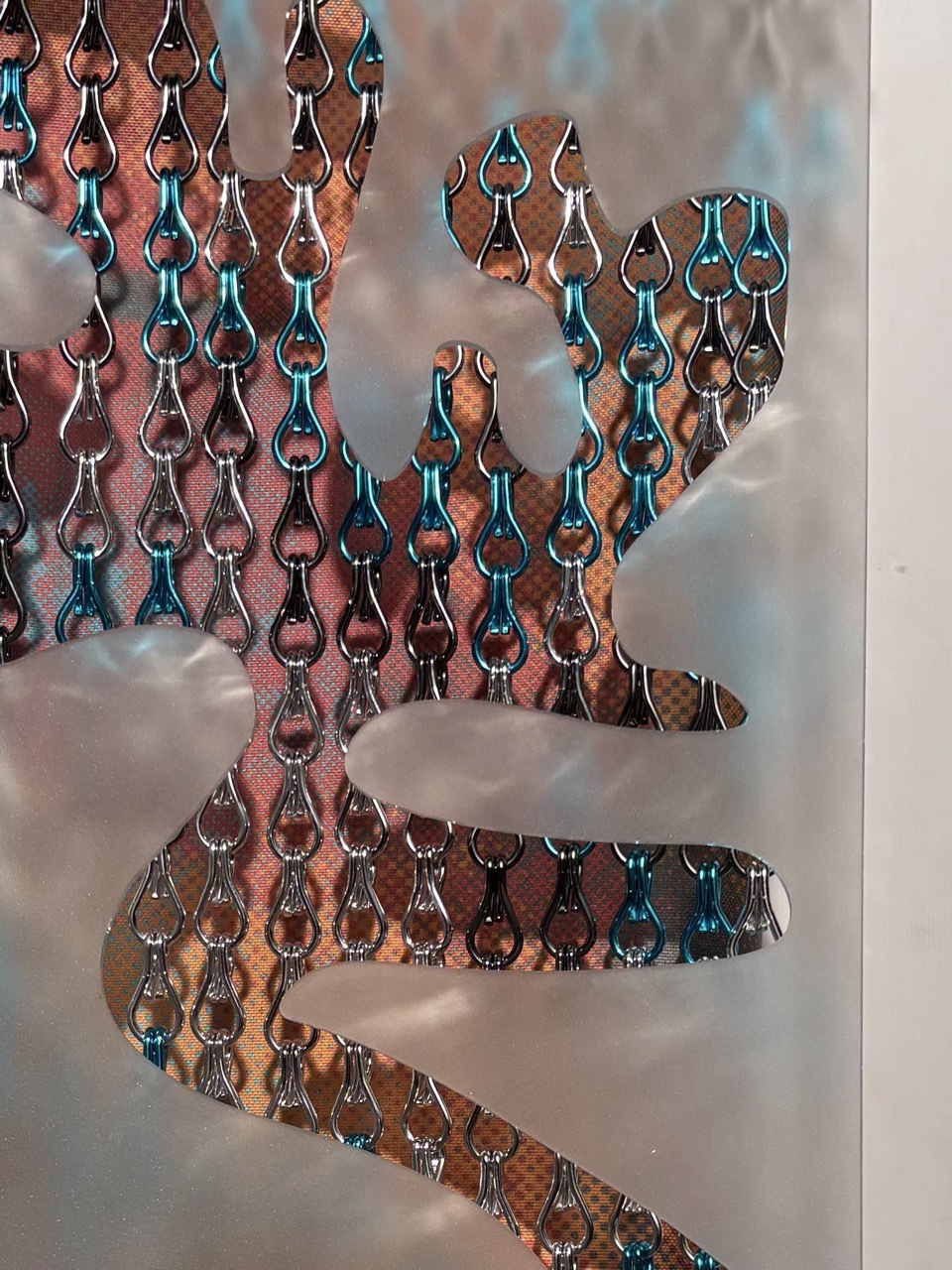 Metallic chainmail curtain with interlocking rings in copper, teal and bronze tones, hanging in curved strips against blurred background.