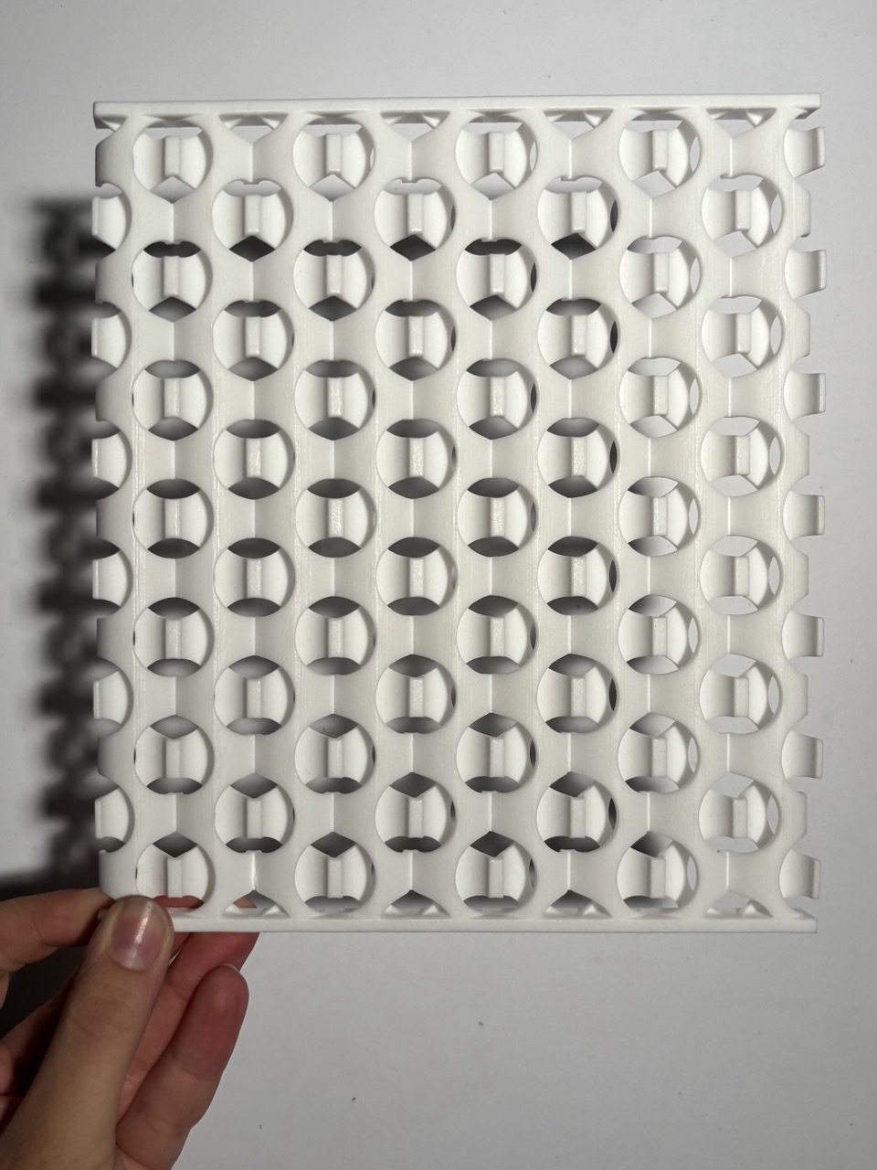 Hand holding white perforated panel with repeating circular pattern creating geometric shadows against white wall.