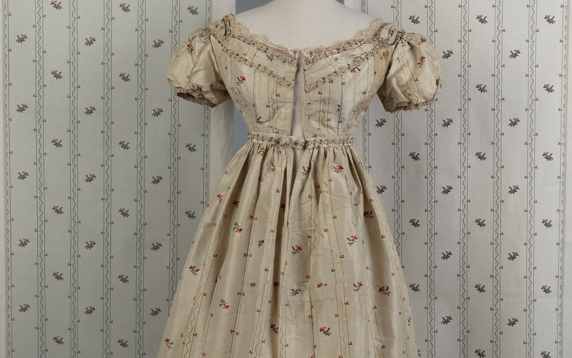 A period-style patterned dress on display in front of similar-patterned wallpaper.