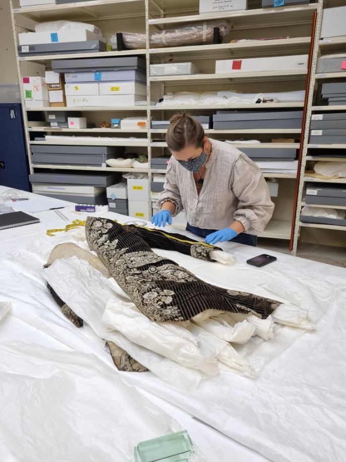 A person wearing a face mask and gloves, standing at a very large table with a historical garment laid out across it. The person is examining one of this garment's sleeves.