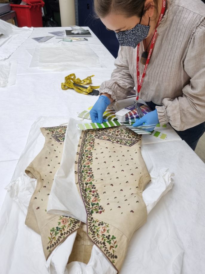 A person in a face mask and gloves holds a series of colour charts next to an embroidered period-style waistcoat, which is laid out on a large table.