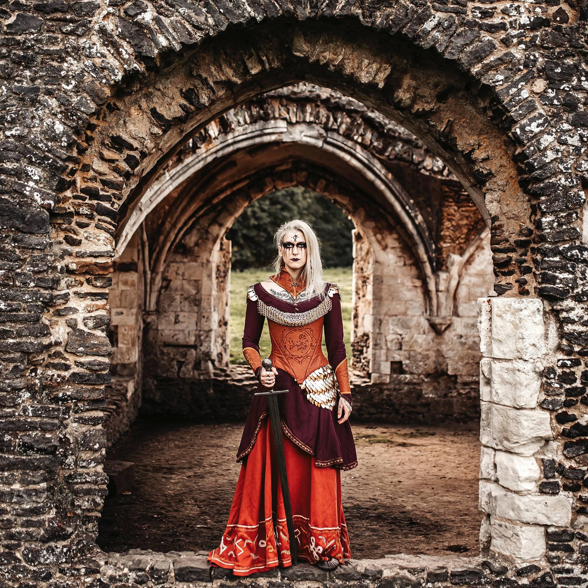A person in Celtic-style dress and make-up stands holding a sword under a set of stone arches.