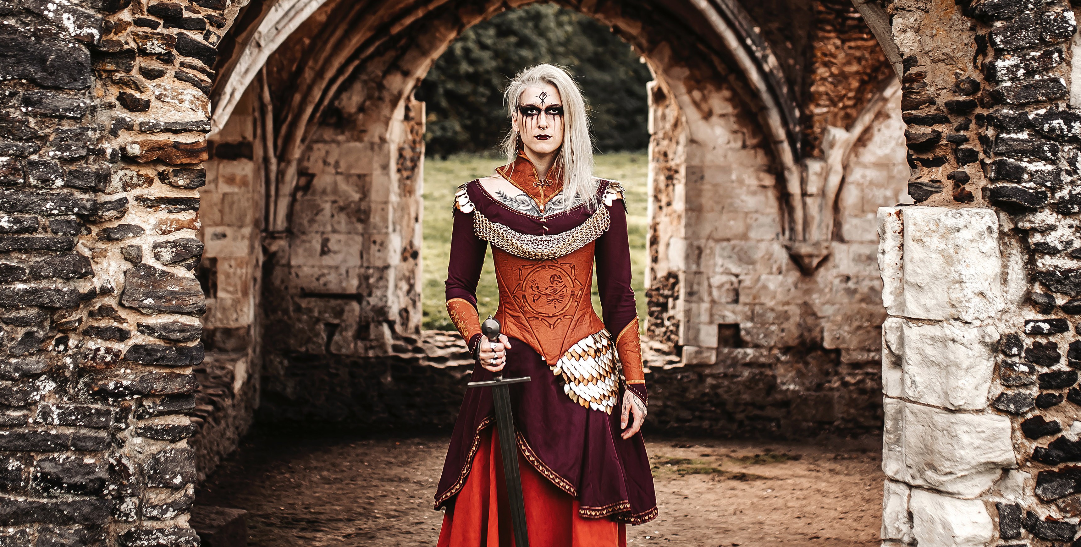 Closer image of a person in Celtic-style dress and make-up stands holding a sword under a set of stone arches.