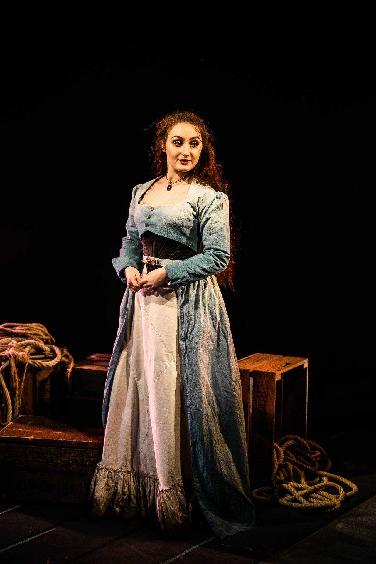 A person in historical-style costume stands on a stage with crates and ropes behind them.