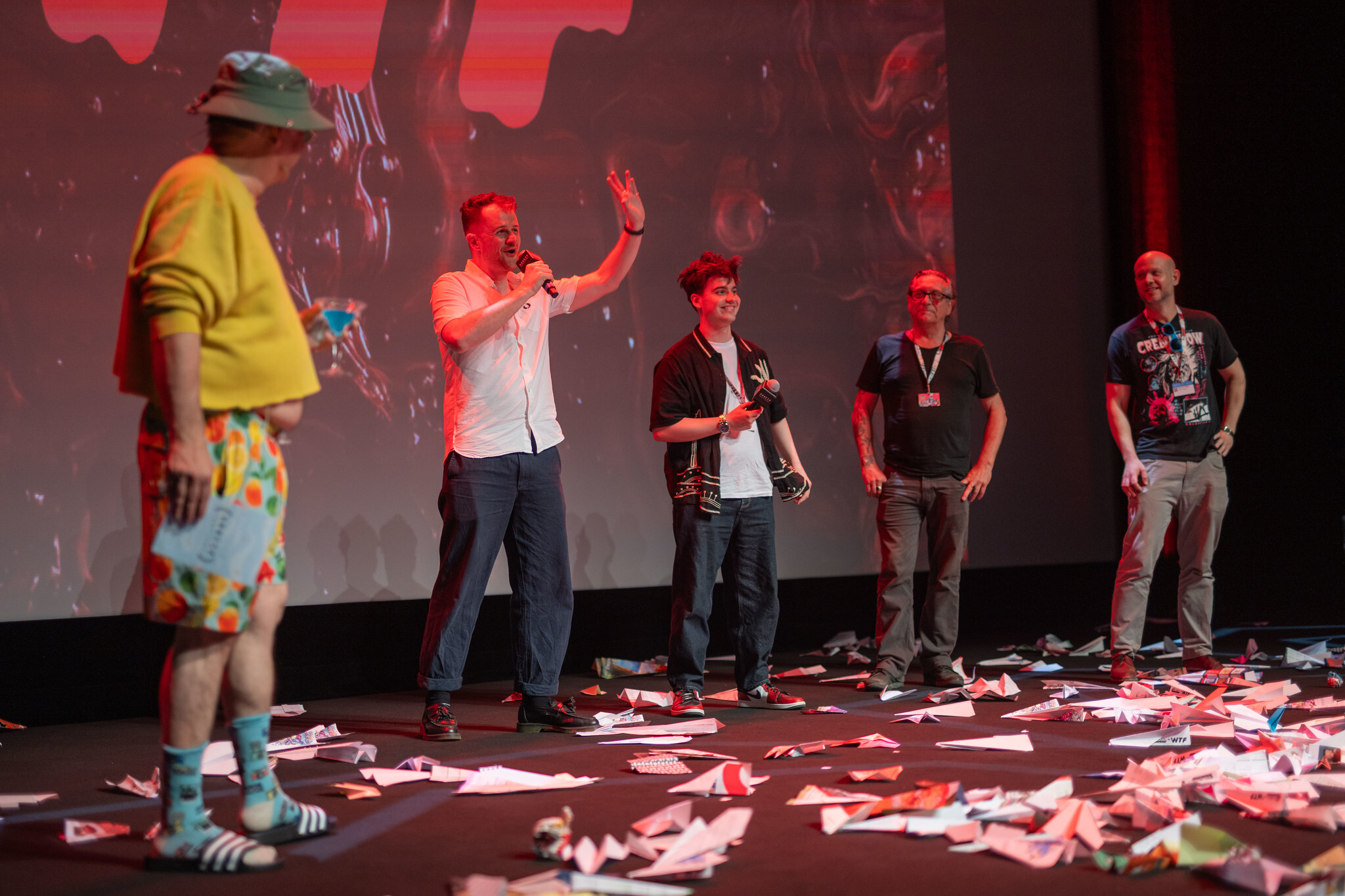 Five people on stage with red lighting, scattered papers on floor, and projection screen behind them showing red abstract shapes.