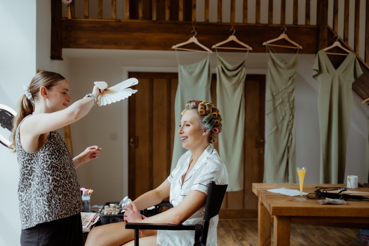 Harmony Evans spraying a woman's hair in rustic room with exposed wooden beams and green garments hanging from banisters..