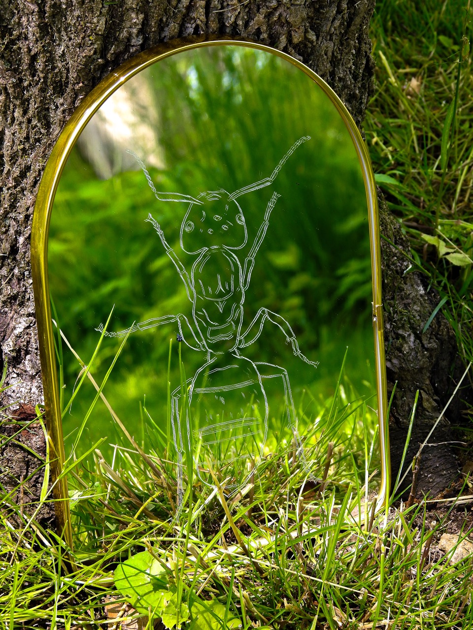 An arched gold-framed mirror etched with an ant stands among grass against a tree trunk.