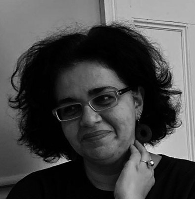 Woman with curly dark hair wearing glasses and dark clothing, smiling at camera in black and white portrait.