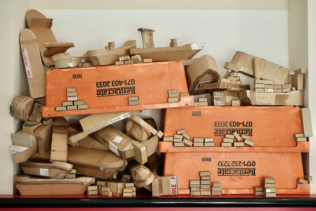 Artwork made up of assorted stacked cardboard boxes and three orange plastic containers.