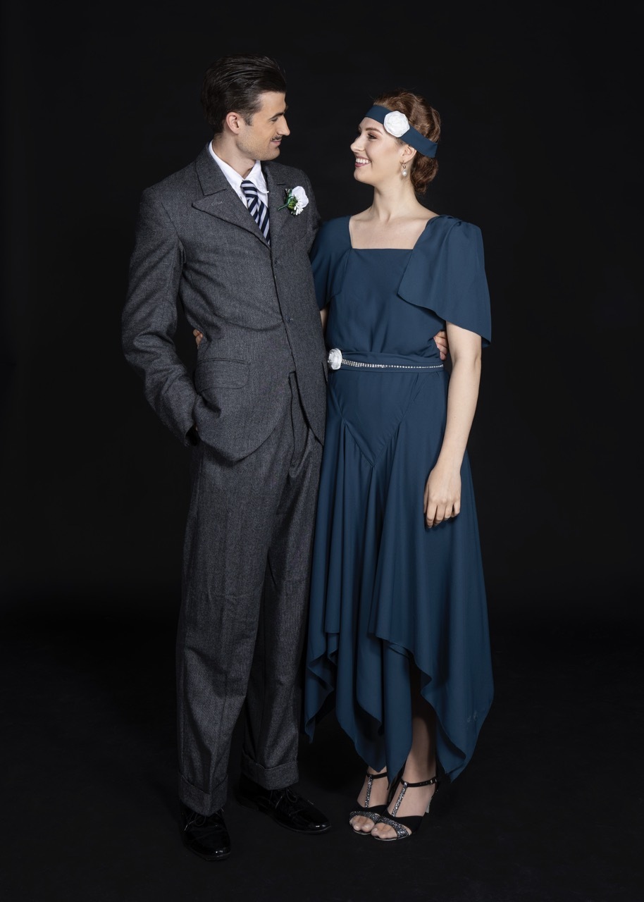 Man in grey three-piece suit standing beside woman in blue dress with white headband, both smiling.