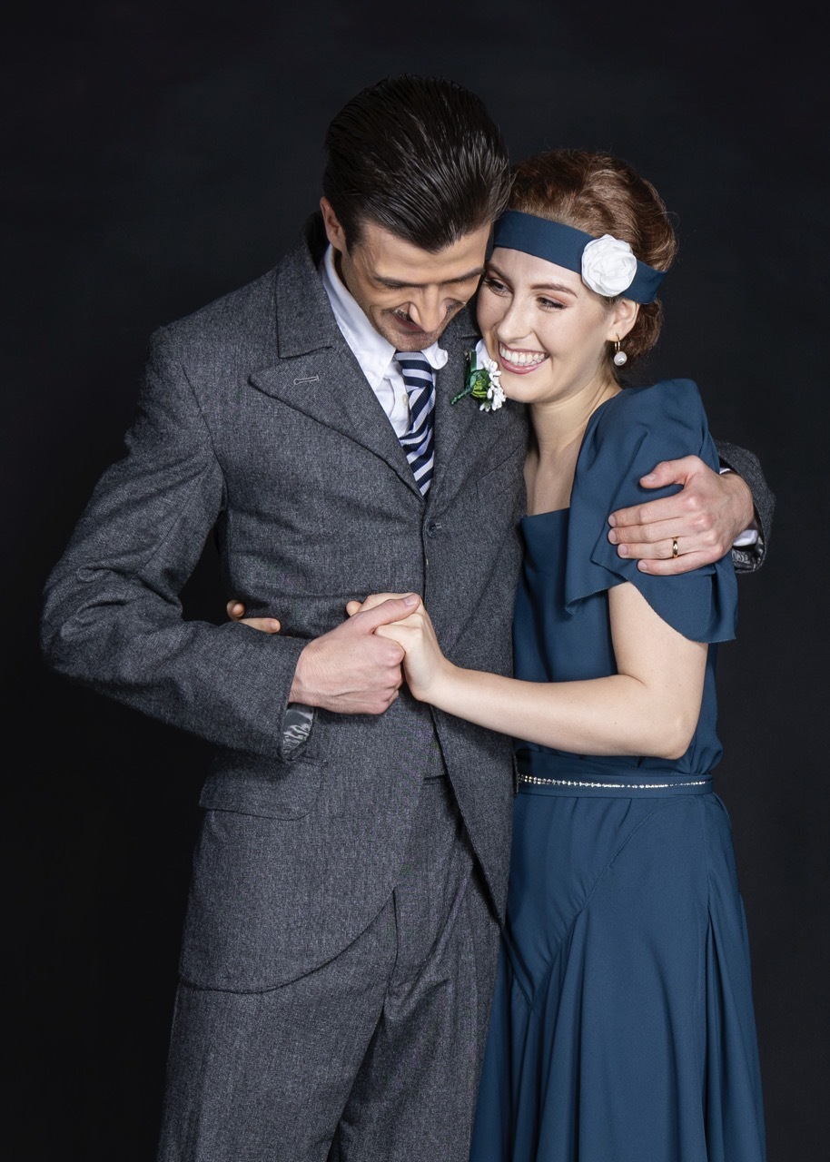 Man in grey suit with striped tie embracing smiling woman in blue dress with white flower headband against dark background.