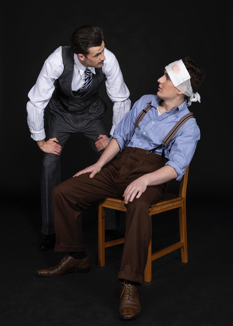 Two men in period costume on black background. One standing in white shirt, black waistcoat and tie, one seated on wooden chair wearing blue shirt, brown braces and white headband plaster, dark background.
