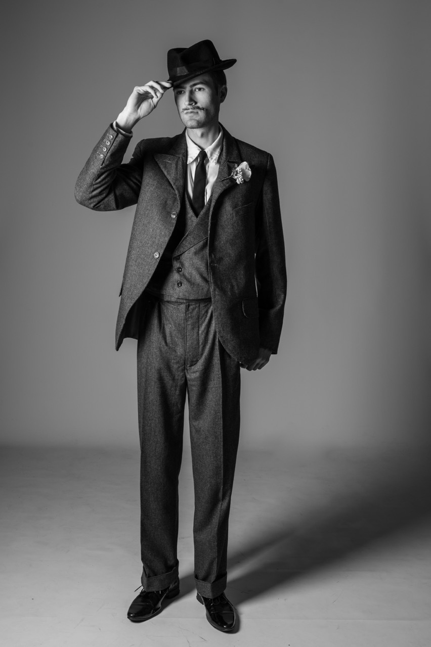 Black and white portrait of man in three-piece suit tipping fedora hat, standing against gradient background.