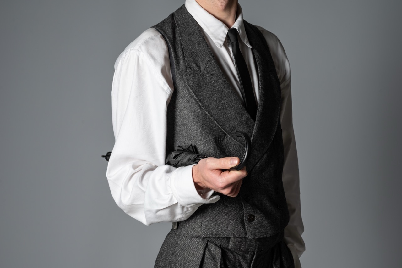 Man wearing grey three-piece waistcoat suit with white shirt and black tie against neutral grey background.