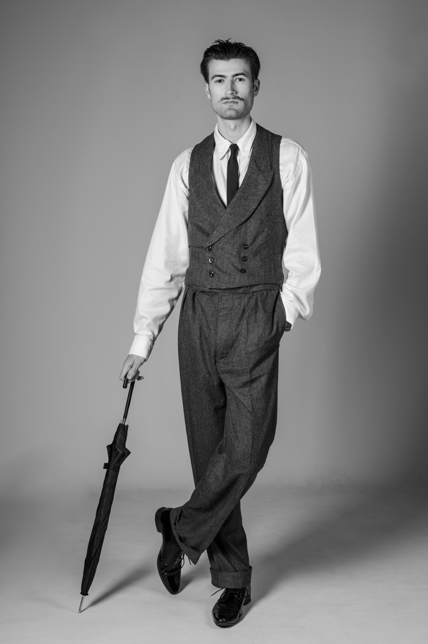Man in vintage waistcoat, white shirt, and dark trousers holds umbrella in black and white portrait against grey backdrop.