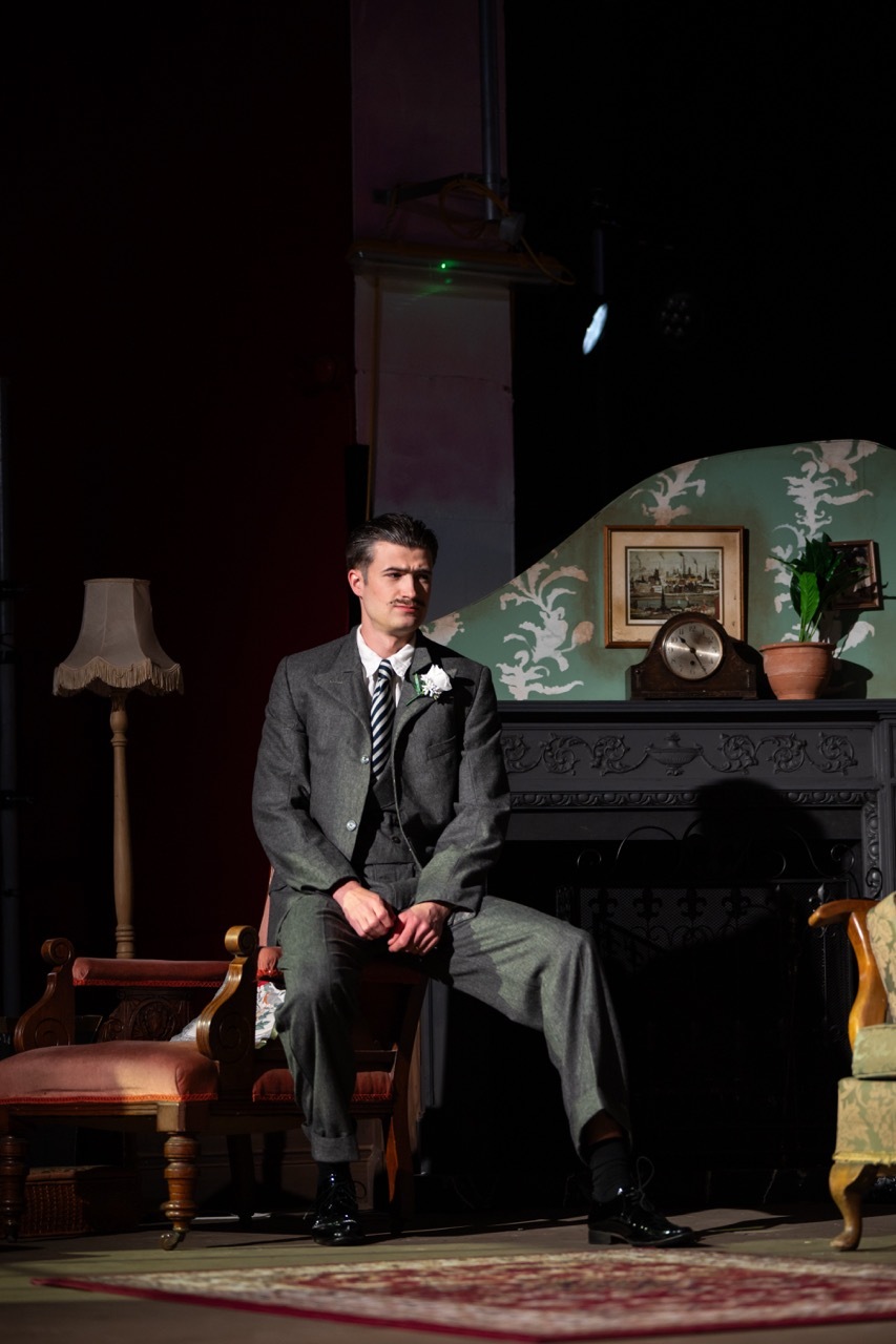 Man in grey suit and striped tie sits on stage set with fireplace, lamp, wooden furniture and patterned rug in theatrical production.