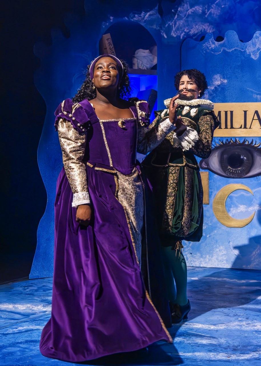 Two theatre performers on stage in period costumes - woman in purple gown with gold trim, man in dark ornate outfit with ruff collar.