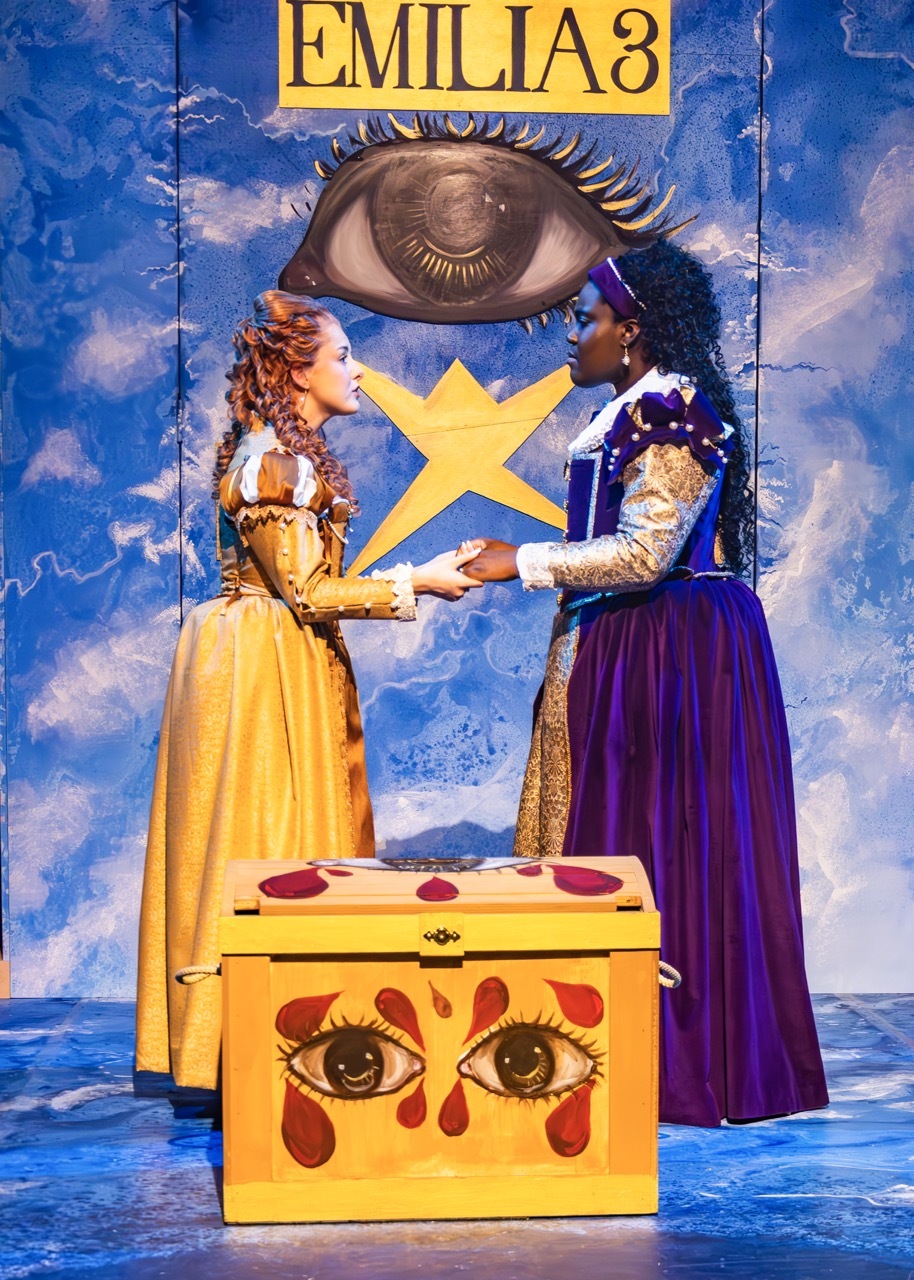 Theatre poster with blue cloudy background. Two women in period dress – one yellow gown, one purple - hold hands above yellow box with painted crying eyes. Large eye and yellow star above. "EMILIA3" text at top.