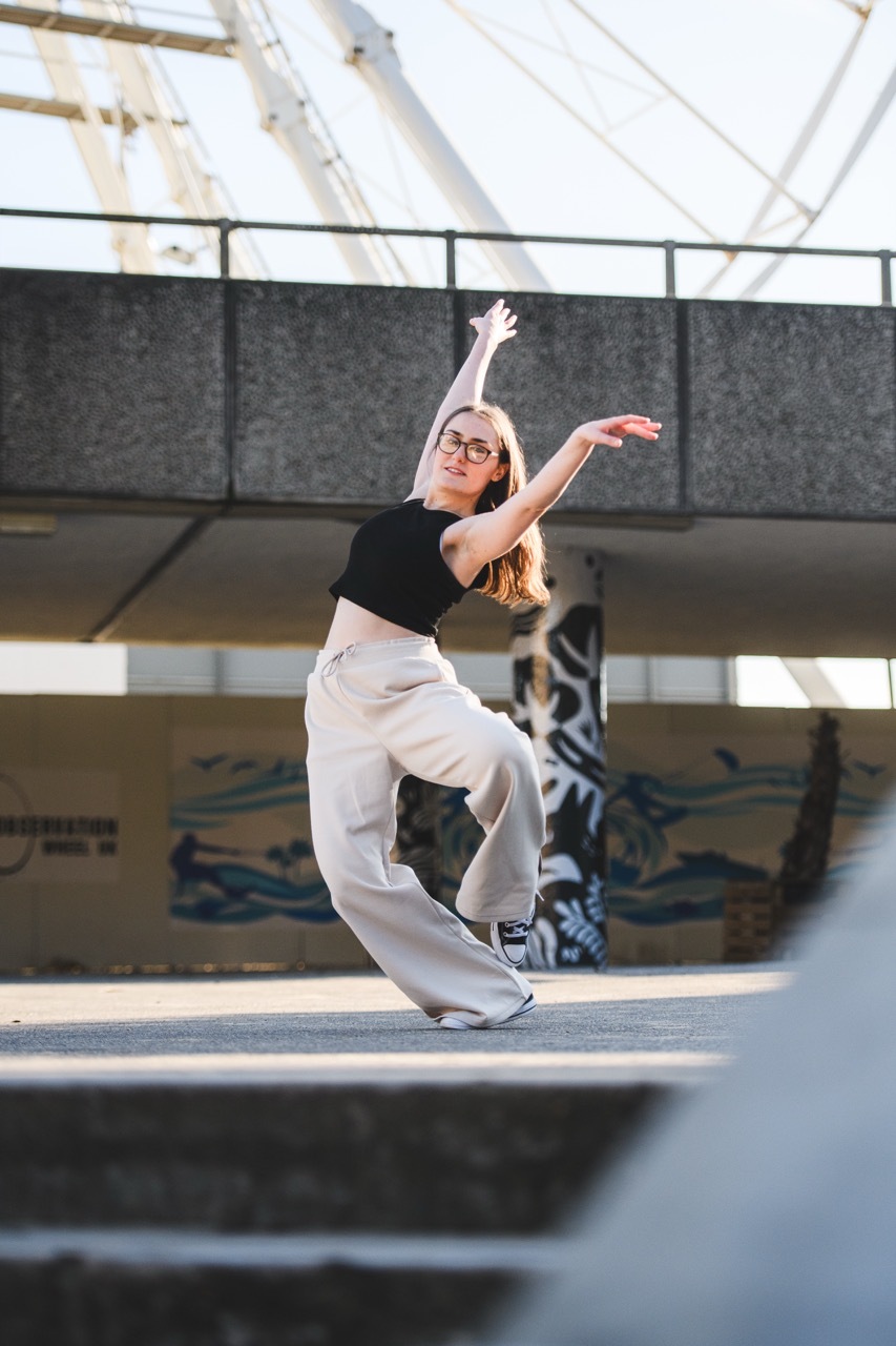Woman in black top and beige trousers dancing with arms extended upward in urban skate park with concrete ramps and metal railings.
