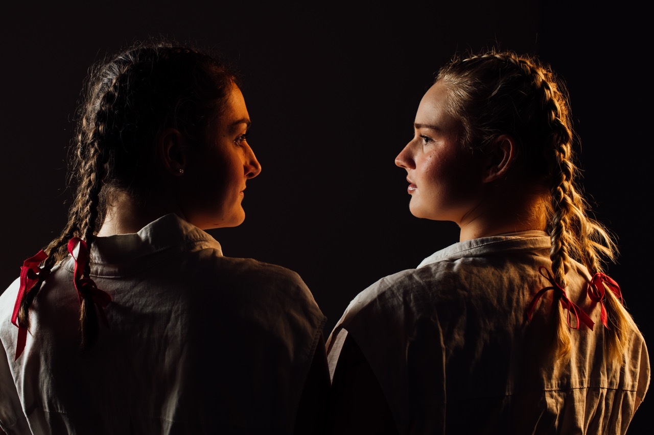 Two actors in profile facing each other against black background, lit with warm orange light, wearing period costumes.