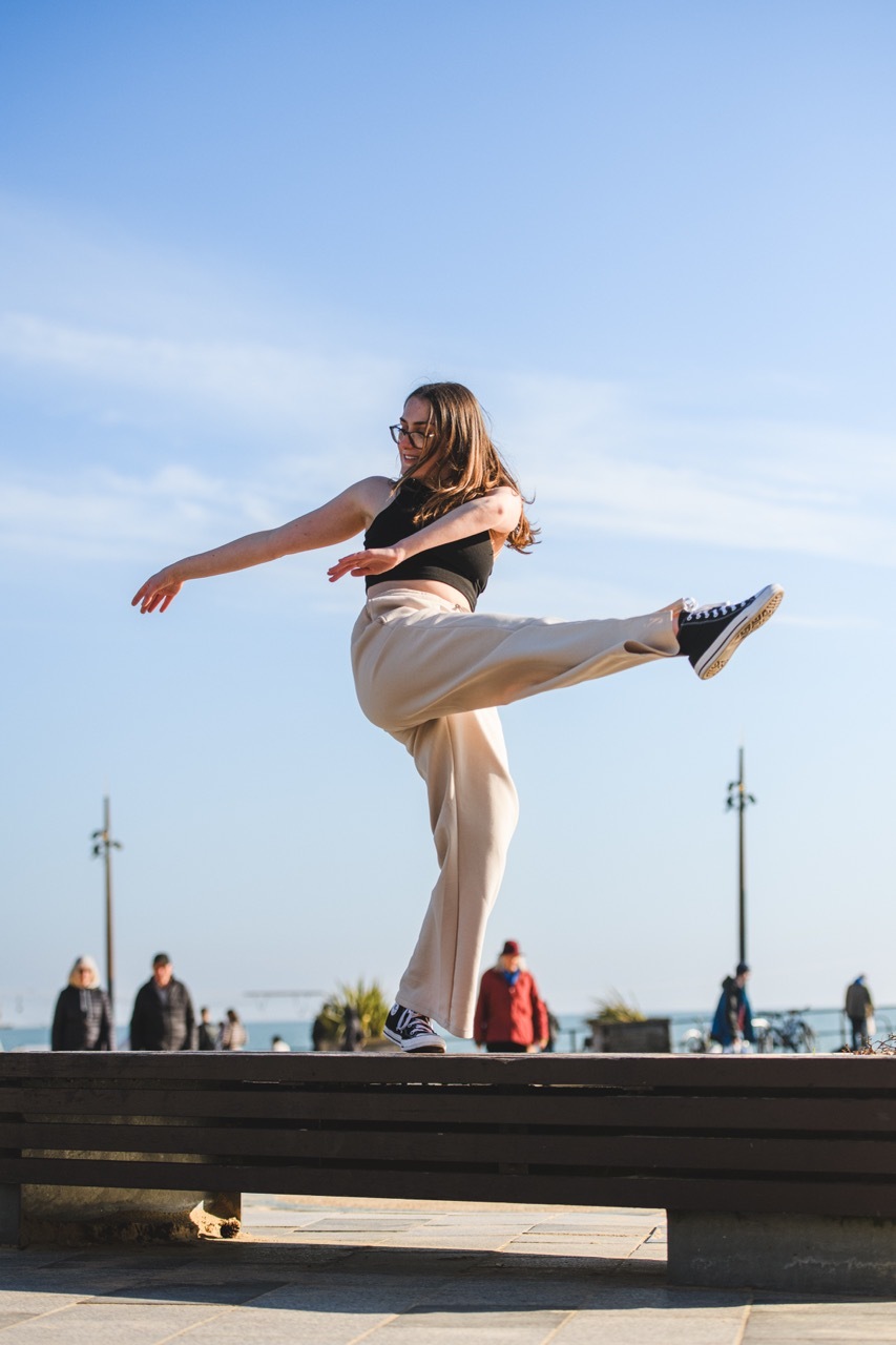 Woman in beige trousers and dark top performing high kick on concrete ledge against blue sky with white clouds and lamp posts.