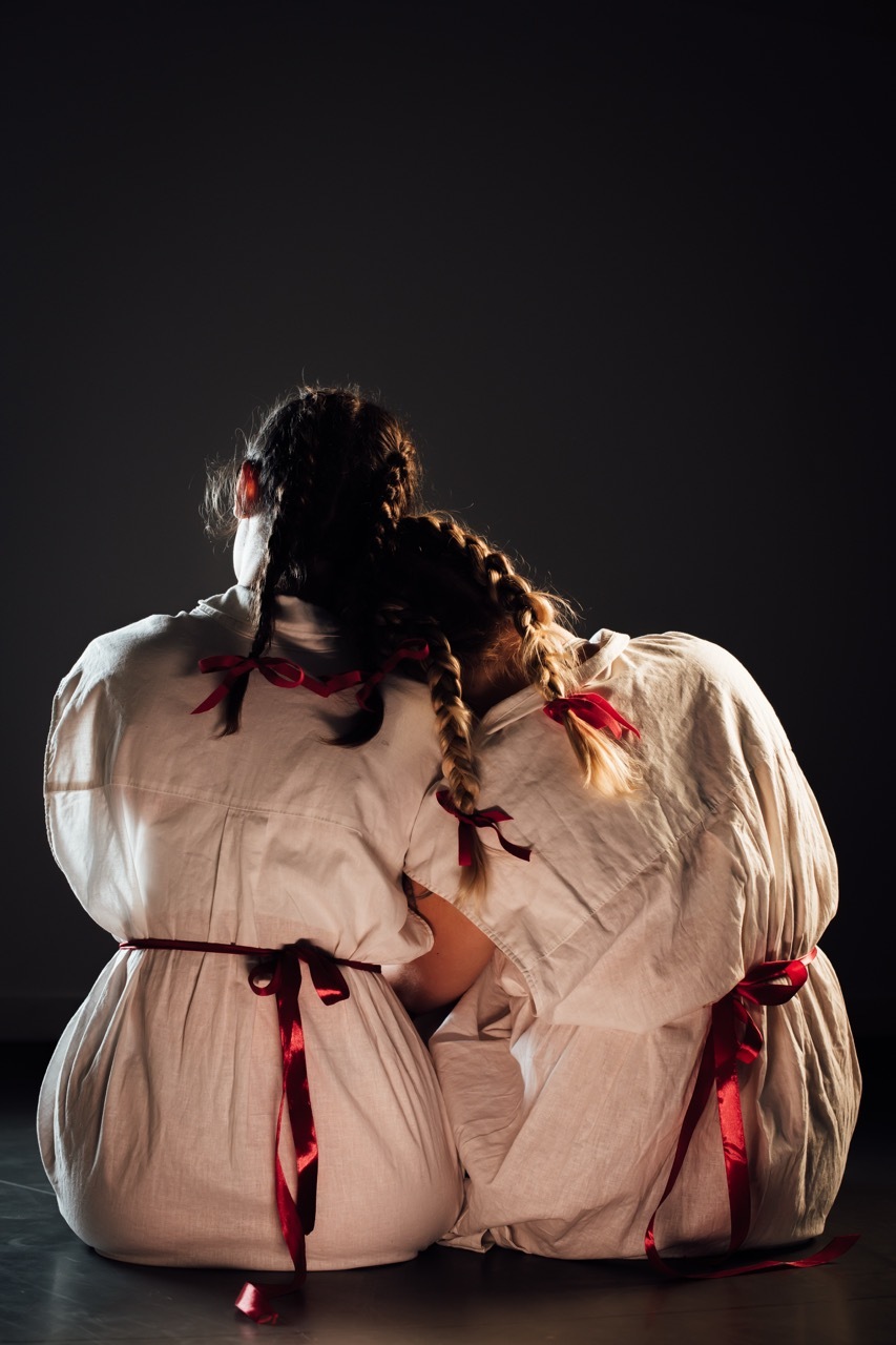 Person in white garment with red ribbons, sitting with head in hands against dark background, dramatic lighting from side.