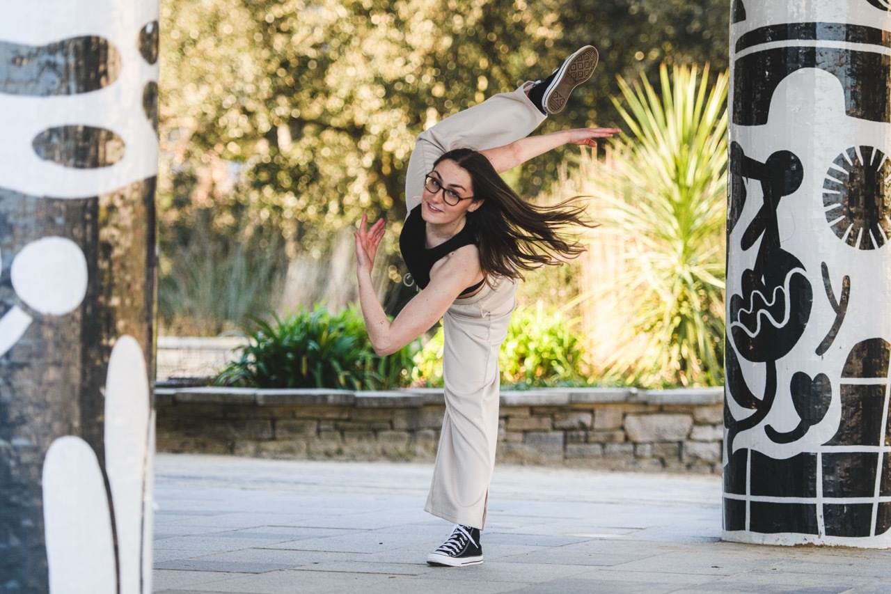 Woman in beige dress and black trainers mid-jump with hair flowing, next to graffitied pillar in urban courtyard with plants.
