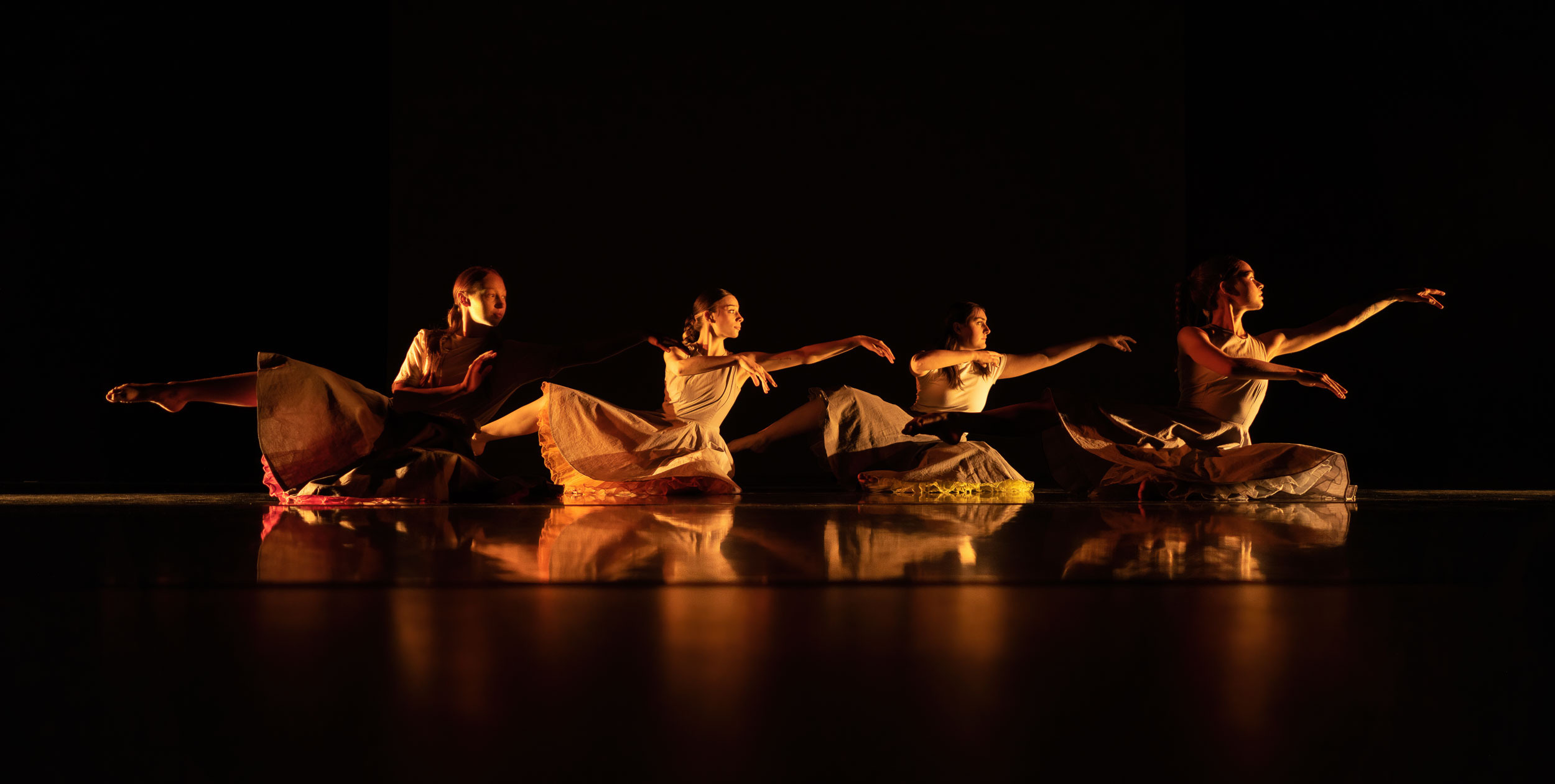 Four dancers in earth-toned costumes perform synchronized movements on a reflective stage floor against a black backdrop, lit by warm amber lighting.