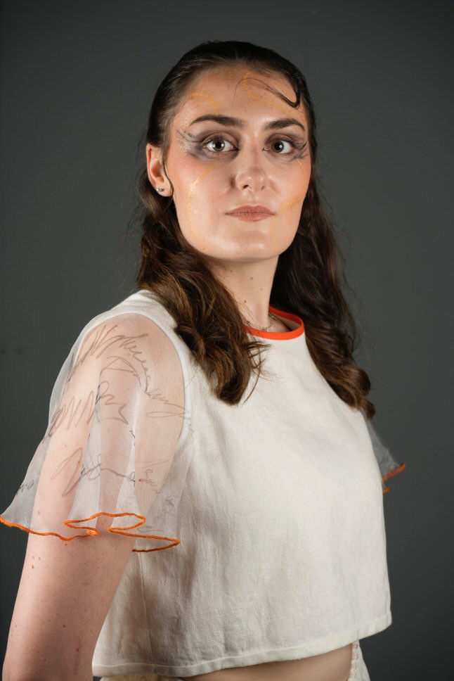 Woman with curly brown hair wearing white top with orange trim, dramatic eye makeup, against grey background.