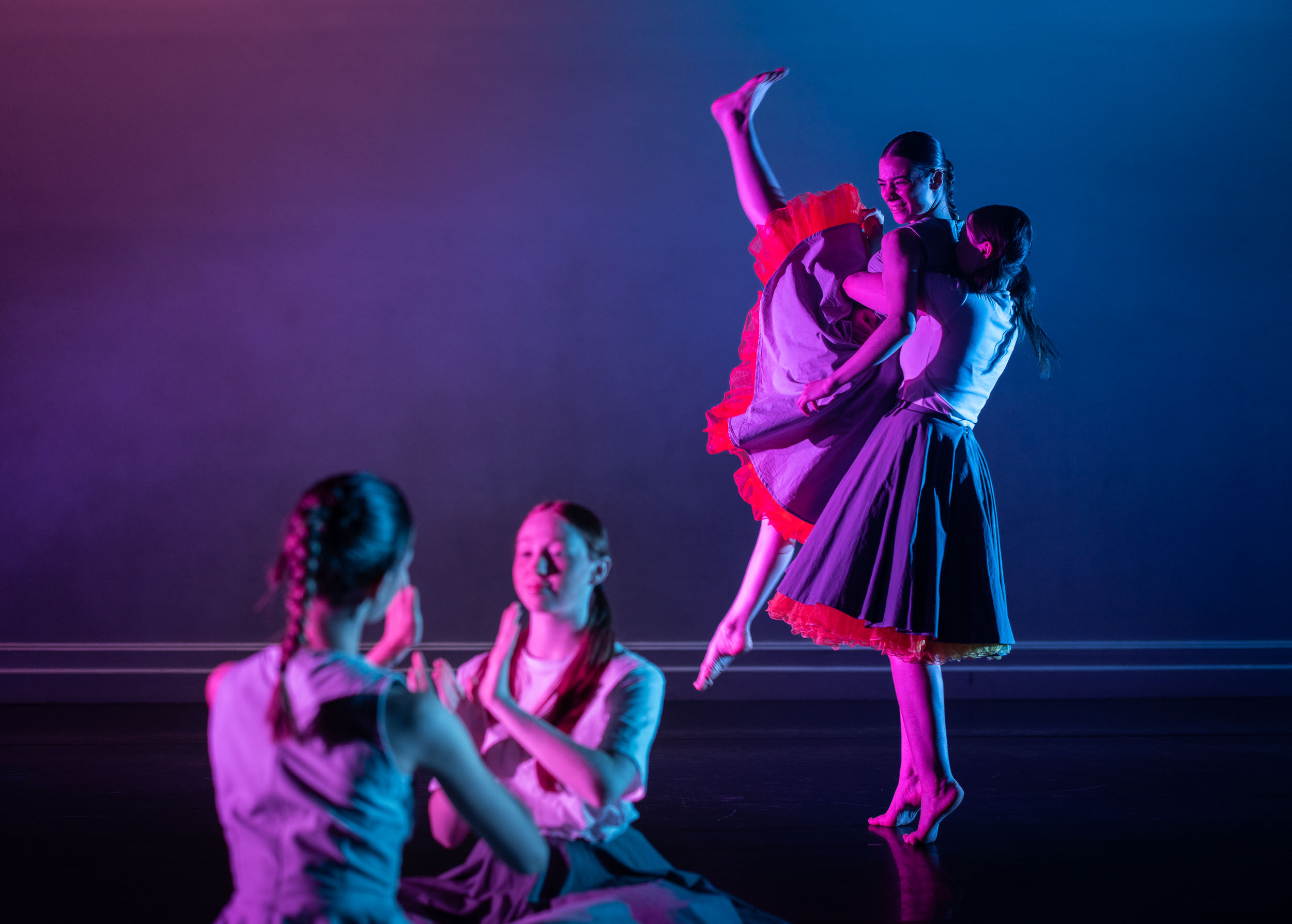 Four dancers in black attire against purple and blue gradient backdrop. Two seated on floor, two standing with raised arms in theatrical pose.