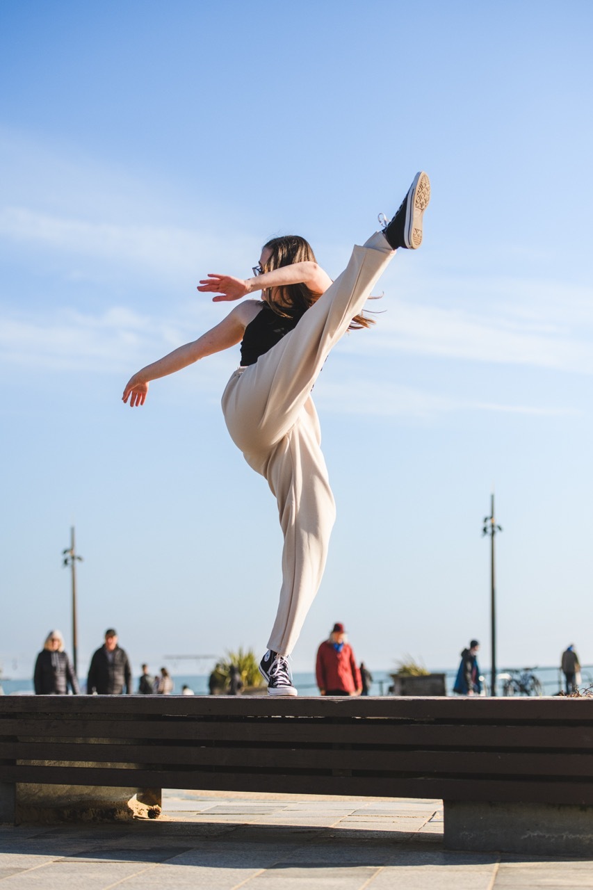 Person in white trousers and black top performing high kick on waterfront promenade with blue sky and distant figures.