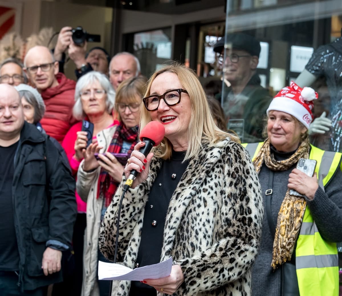 A. Pearson speaks into microphone whilst crowd watches behind her on street.