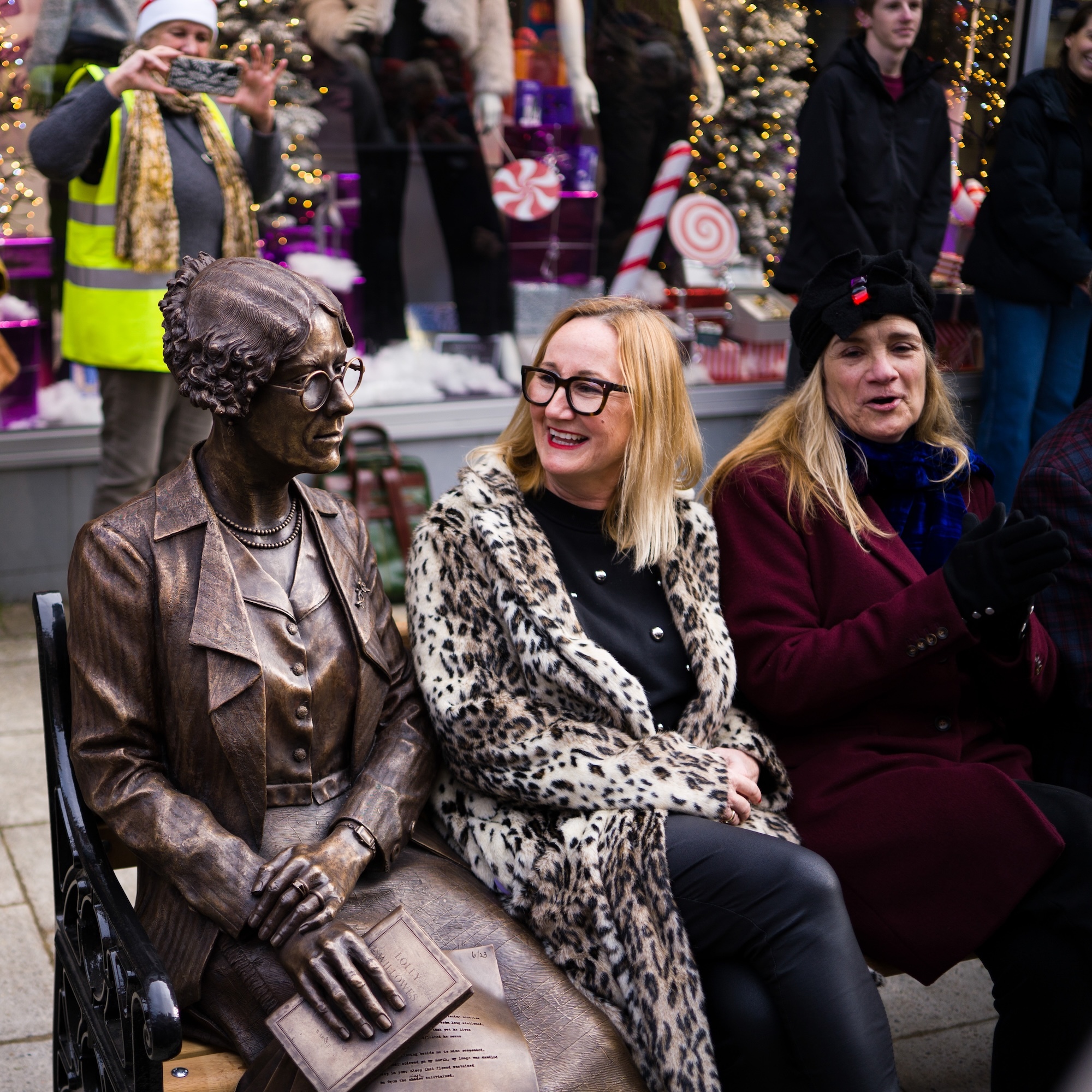 Two people sitting on bench with bronze statue of S Townsend Warner in centre. Left person wears leopard print coat, right wears burgundy coat.