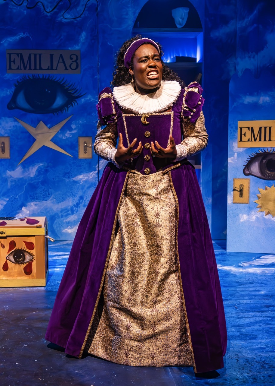 Performer in purple velvet and gold brocade Elizabethan gown with white ruff collar on blue stage set with "EMILIA" signage.