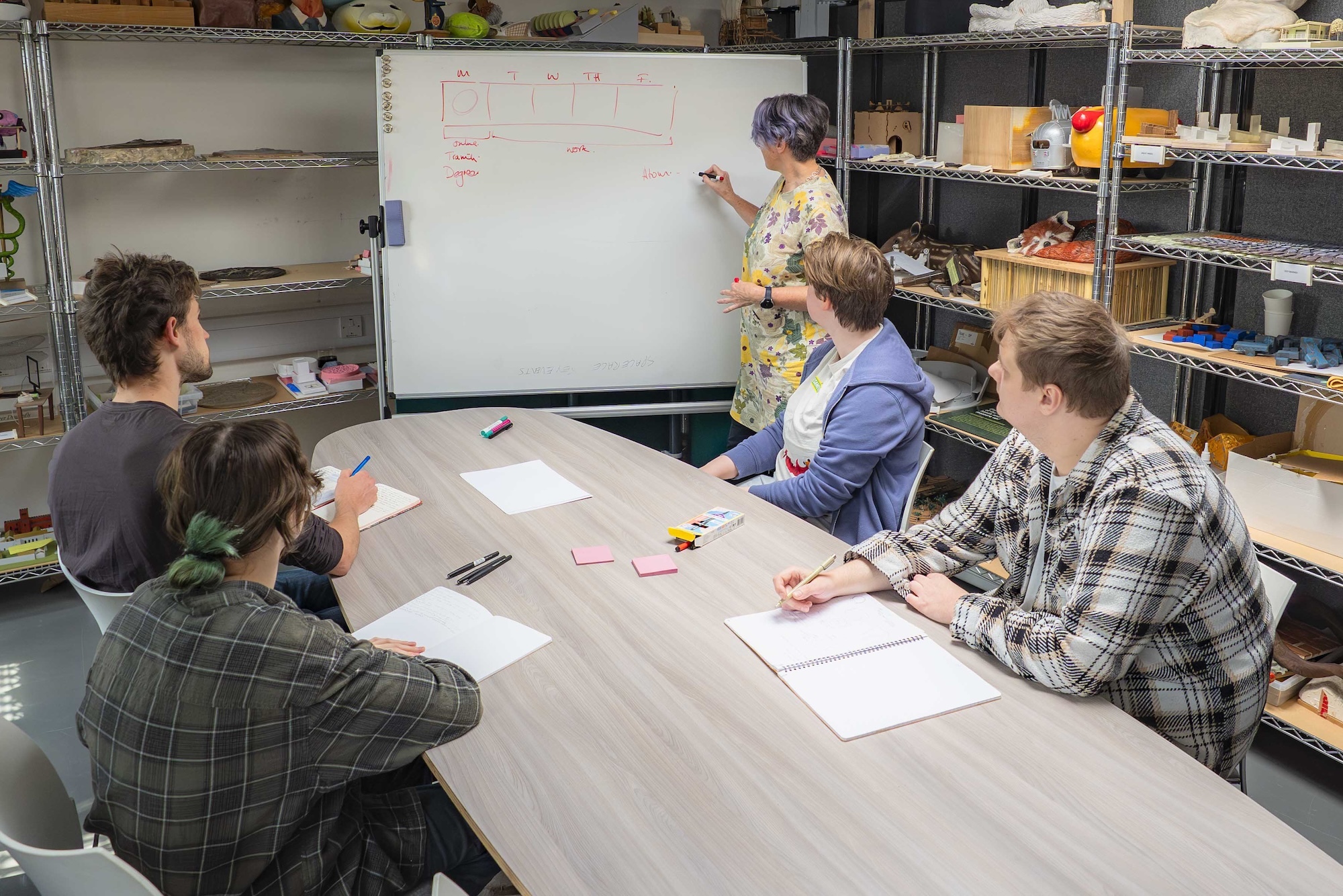 Students seated around table with papers while a lecturer writes on whiteboard in classroom with storage shelves.
