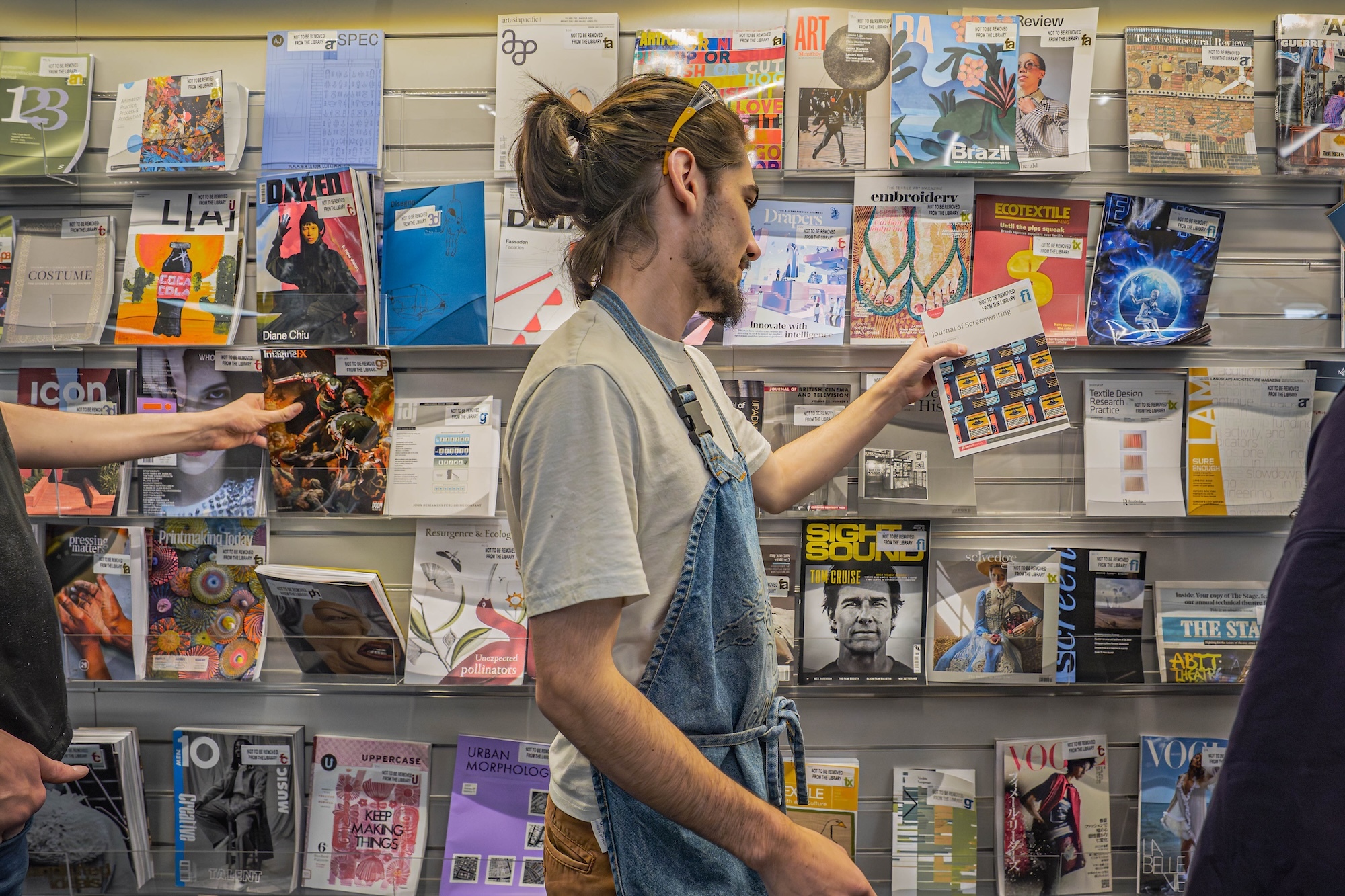 Person with brown hair in bun wearing white t-shirt and denim dungarees browsing records at shop with colourful album covers on wall display.
