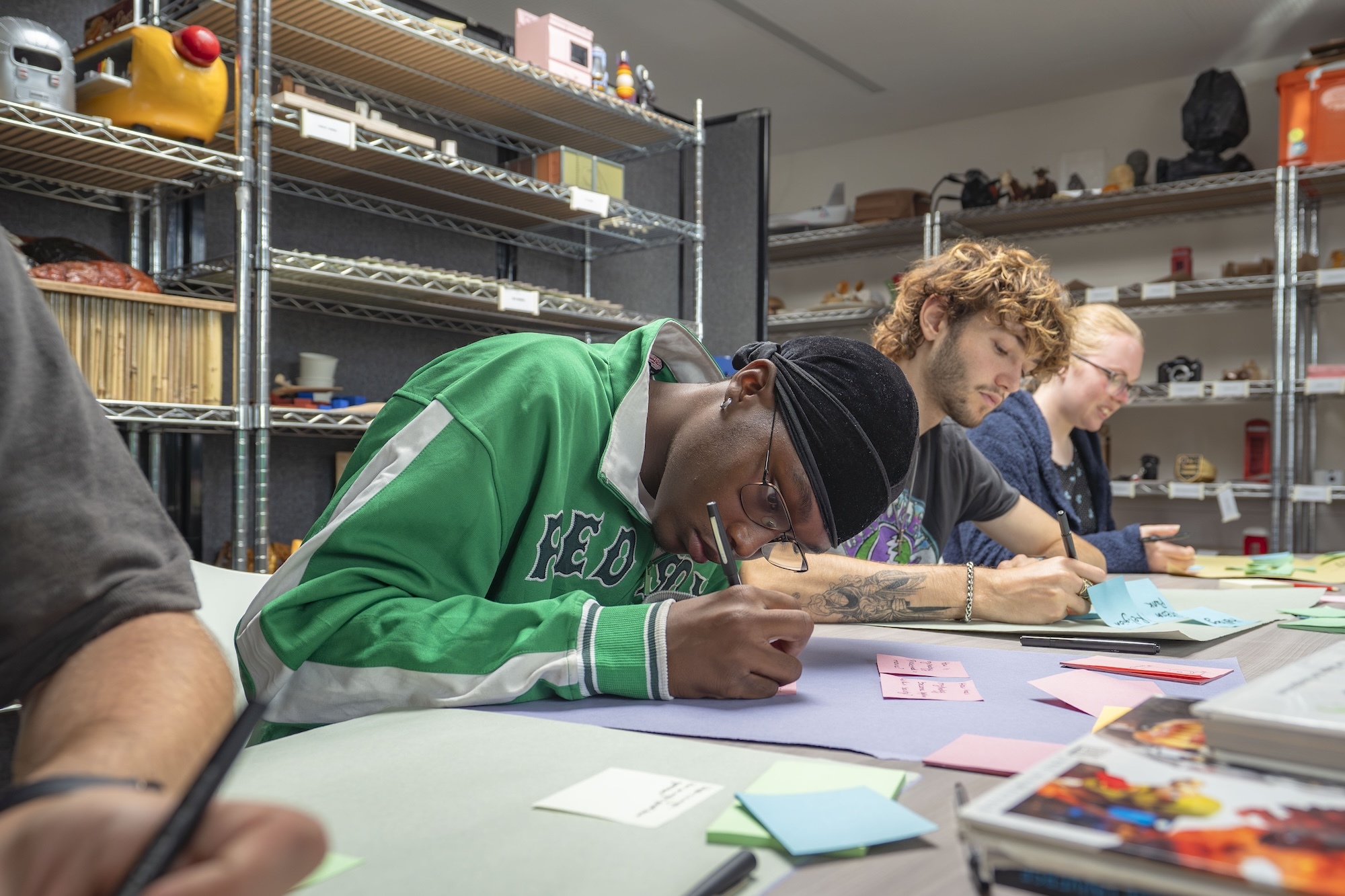 Three people working at table with papers and art supplies, surrounded by metal shelving units filled with materials and equipment.