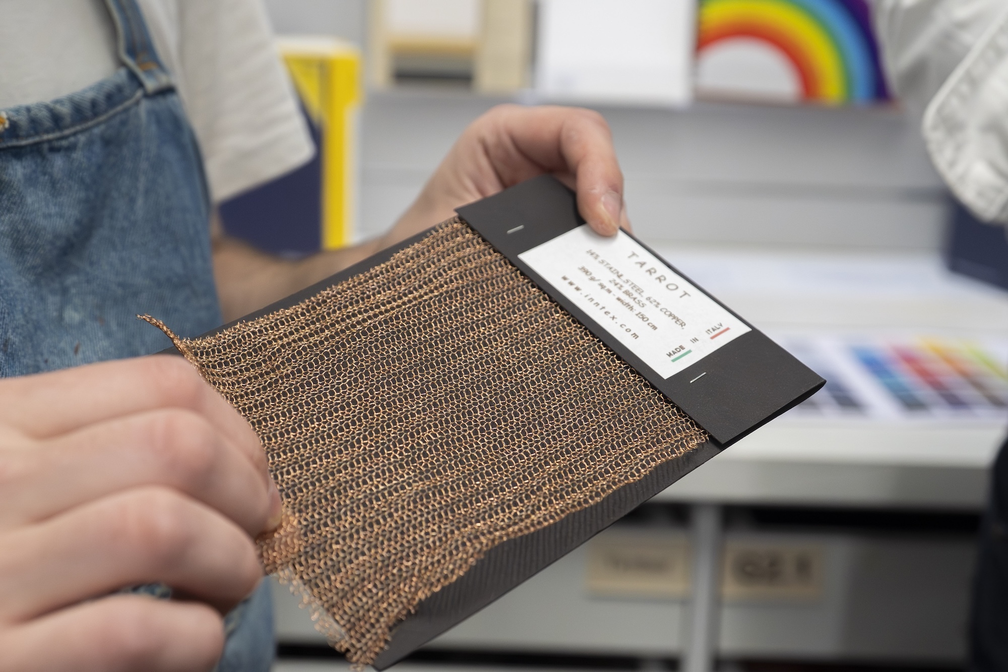 Person holding brown corrugated cardboard sample with white label, classroom setting with rainbow decoration in background.