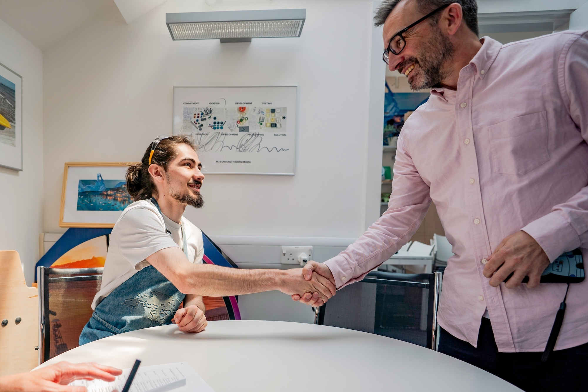 Two men shaking hands in modern office with colourful artwork on white walls, one seated at desk, one standing wearing glasses.