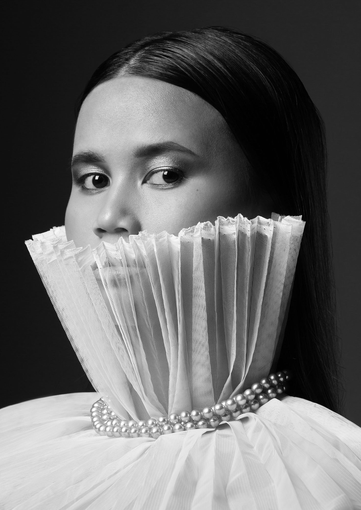 Black and white headshot of a model with a high upturned collar going above their mouth, and strings of pearls around their neck.