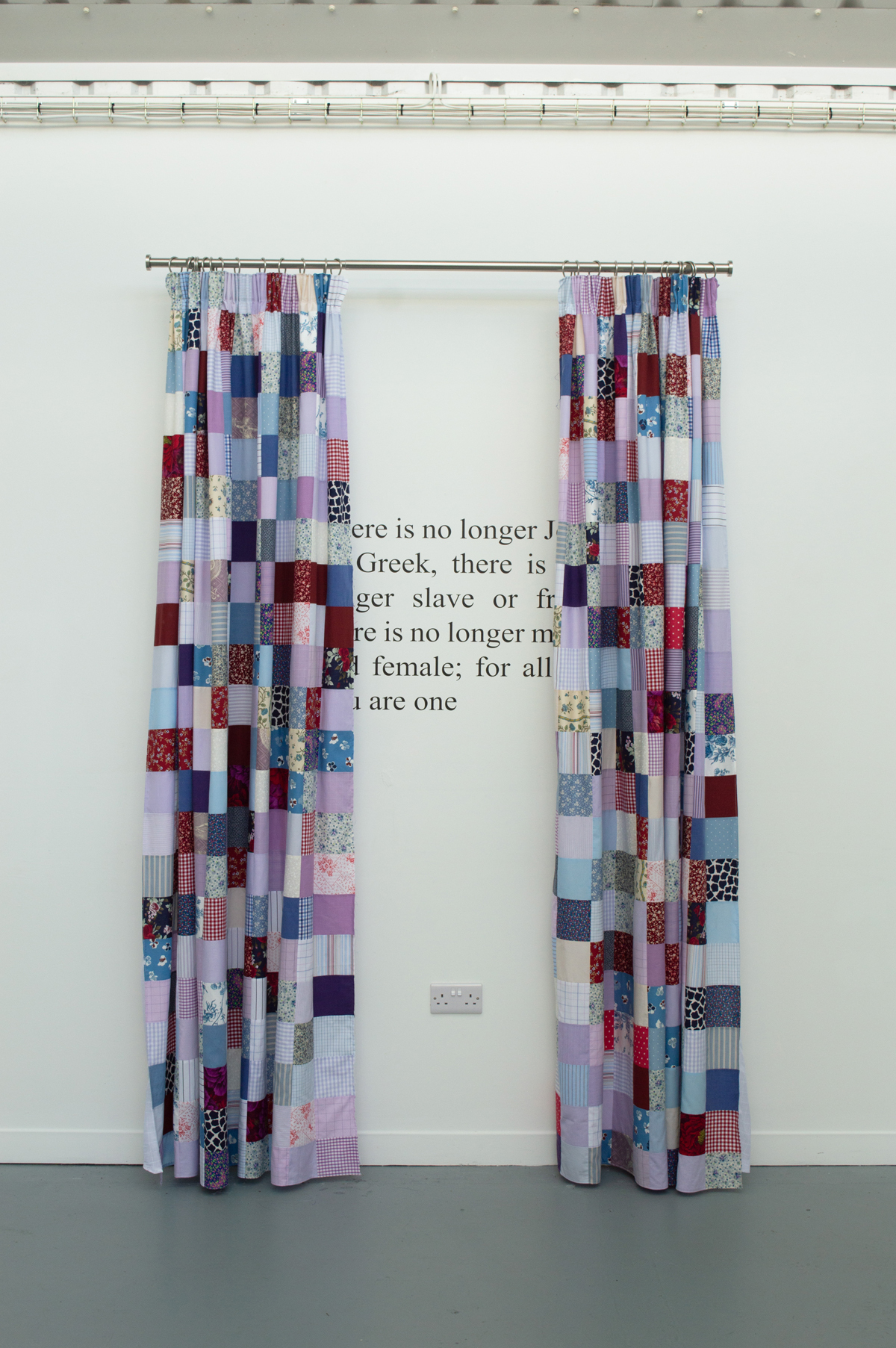Patchwork curtains made of multicoloured fabric squares hang on white wall with partial text about Greek identity visible between panels.